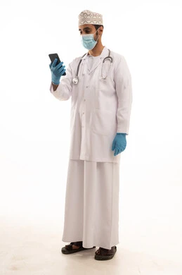 اتباع اجراءات السلامة للوقاية من فايروس كورونا، دمج التقنية والتكنولوجيا في الطب والرعاية الصحية، العمل في القطاع الطبي في سلطنة عمان، بورتريه لطبيب عربي خليجي عماني يرتدي الدشداشة والكمة والزي الخاص بالمهنة يضع السماعة الطبية يستخدم  الهاتف المحمول بتعابير السرور، الجسم بالطول الكامل، خلفية بيضاء