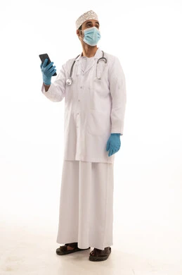 اتباع اجراءات السلامة للوقاية من فايروس كورونا، دمج التقنية والتكنولوجيا في الطب والرعاية الصحية، العمل في القطاع الطبي في سلطنة عمان، بورتريه لطبيب عربي خليجي عماني يرتدي الدشداشة والكمة والزي الخاص بالمهنة يضع السماعة الطبية يستخدم  الهاتف المحمول بتعابير السرور، الجسم بالطول الكامل، خلفية بيضاء
