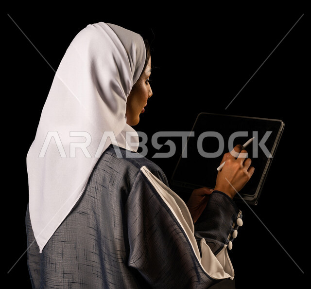 صورة بزاوية لسيدة عربية خليجية سعودية تمسك جهاز تقني تابلت بإيماءات و تعابير وجه و يدين مختلفة ، سيدة أعمال ، تدوين الملاحظات و مراجعتها ، استخدام الأجهزة التقنية في العمل ، خلفية سوداء