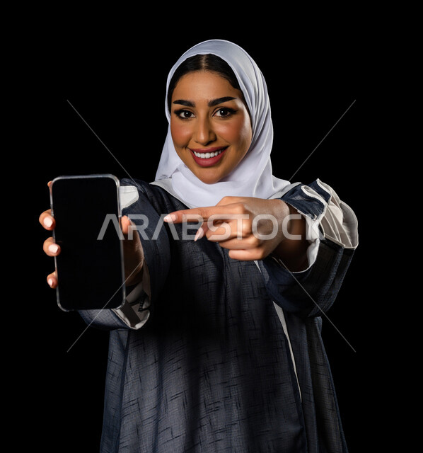 بورتريه لسيدة عربية خليجية سعودية مبتسمة تحمل بيدها الهاتف المحمول و تشير بأصبع السبابة اتجاه الجهاز، التسويق الرقمي ، اعلانات تسويقية ، تطبيقات الهاتف الذكي ، خلفية سوداء