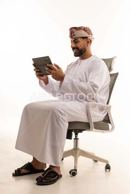 رجل عُماني يستخدم جهاز لوحي على كرسي مكتب بخلفية بيضاء