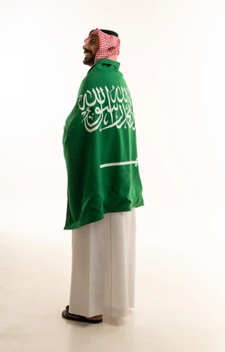 رجل سعودي يرتدي الزي التقليدي وملتف بالعلم الوطني