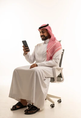 رجل سعودي بالثوب والشماغ يستخدم الهاتف الذكي على كرسي