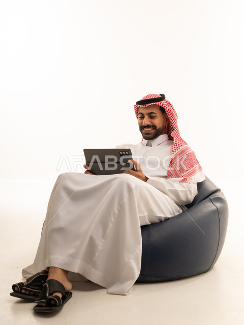 رجل سعودي يستخدم جهازاً لوحياً على مقعد مريح خلفية بيضاء