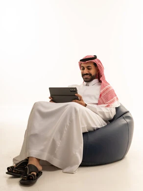 رجل سعودي يستخدم جهازاً لوحياً على مقعد مريح خلفية بيضاء