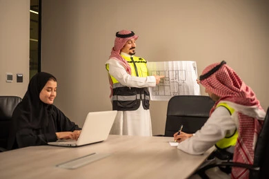 دراسة الرسوم البيانية والاحصاءات، متابعة مخطط الاعمار, العمل في القطاع الهندسي، مفهوم الهندسة والانشاءات المعمارية، مهندس عربي خليجي سعودي يرتدي الثوب التقليدي والشماغ وسترة الحماية يمسك بيده المخطط يجلس مع موظفة تمسك بيدها جهاز اللابتوب ومهندس معماري  ، مهنة ووظيفة في السعودية