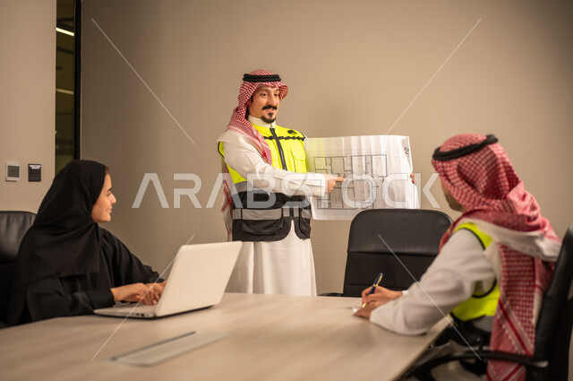 دراسة الرسوم البيانية والاحصاءات، متابعة مخطط الاعمار, العمل في القطاع الهندسي، مفهوم الهندسة والانشاءات المعمارية، مهندس عربي خليجي سعودي يرتدي الثوب التقليدي والشماغ وسترة الحماية يمسك بيده المخطط يجلس مع موظفة تمسك بيدها جهاز اللابتوب ومهندس معماري  ، مهنة ووظيفة في السعودية