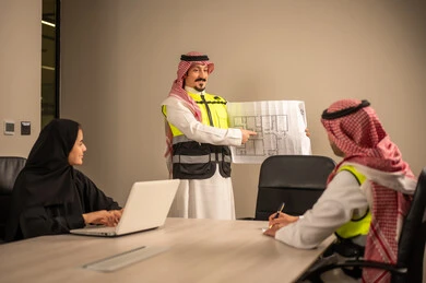 دراسة الرسوم البيانية والاحصاءات، متابعة مخطط الاعمار, العمل في القطاع الهندسي، مفهوم الهندسة والانشاءات المعمارية، مهندس عربي خليجي سعودي يرتدي الثوب التقليدي والشماغ وسترة الحماية يمسك بيده المخطط يجلس مع موظفة تمسك بيدها جهاز اللابتوب ومهندس معماري  ، مهنة ووظيفة في السعودية