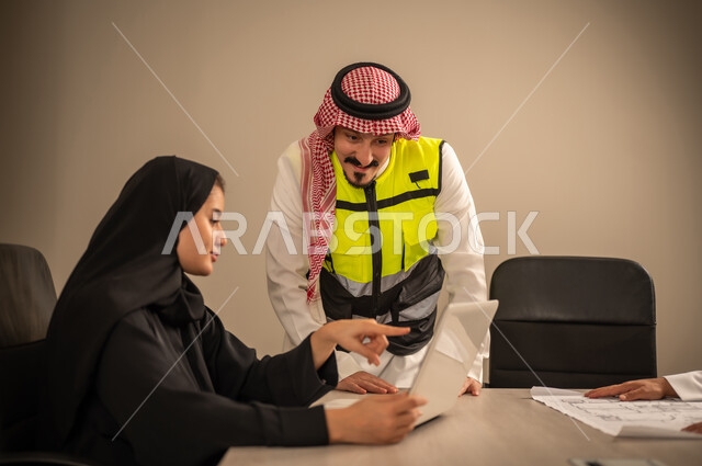 دمج التقنيات المتطورة فى العمل، مفهوم الهندسة والانشاءات المعمارية،  العمل في القطاع الهندسي، متابعة مخطط الاعمار, مهندسة عربية خليجية سعودية ترتدي العباءة والحجاب تستخدم جهاز اللابتوب وتتناقش مع مهندس معماري ، مهنة ووظيفة في السعودية