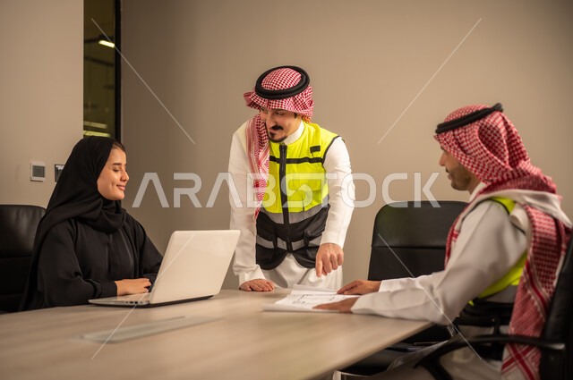 دراسة الرسوم البيانية والاحصاءات، متابعة مخطط الاعمار, العمل في القطاع الهندسي، مفهوم الهندسة والانشاءات المعمارية، مهندس عربي خليجي سعودي يرتدي الثوب التقليدي والشماغ وسترة الحماية يمسك بيده المخطط يجلس مع موظفة تمسك بيدها جهاز اللابتوب ومهندس معماري  ، مهنة ووظيفة في السعودية
