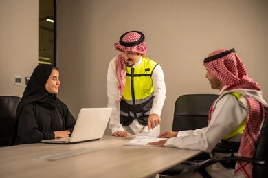 دراسة الرسوم البيانية والاحصاءات، متابعة مخطط الاعمار, العمل في القطاع الهندسي، مفهوم الهندسة والانشاءات المعمارية، مهندس عربي خليجي سعودي يرتدي الثوب التقليدي والشماغ وسترة الحماية يمسك بيده المخطط يجلس مع موظفة تمسك بيدها جهاز اللابتوب ومهندس معماري  ، مهنة ووظيفة في السعودية