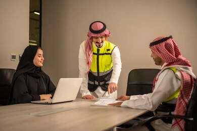 دراسة الرسوم البيانية والاحصاءات، متابعة مخطط الاعمار, العمل في القطاع الهندسي، مفهوم الهندسة والانشاءات المعمارية، مهندس عربي خليجي سعودي يرتدي الثوب التقليدي والشماغ وسترة الحماية يمسك بيده المخطط يجلس مع موظفة تمسك بيدها جهاز اللابتوب ومهندس معماري  ، مهنة ووظيفة في السعودية