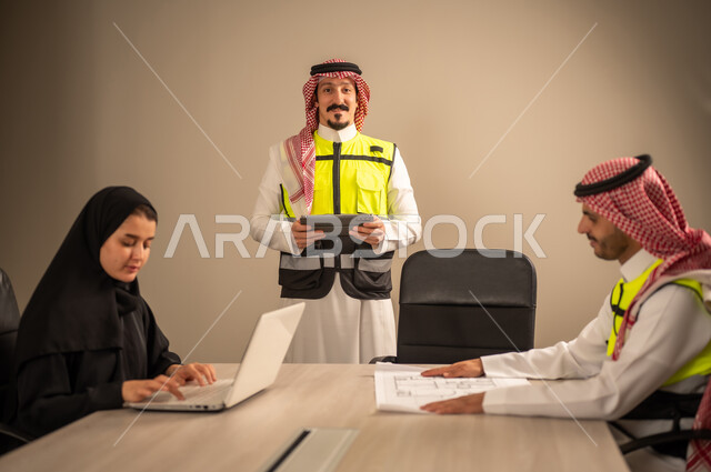 دراسة الرسوم البيانية والاحصاءات، متابعة مخطط الاعمار, العمل في القطاع الهندسي، مفهوم الهندسة والانشاءات المعمارية، مهندس عربي خليجي سعودي يرتدي الثوب التقليدي والشماغ وسترة الحماية يمسك بيده المخطط يجلس مع موظفة تمسك بيدها جهاز اللابتوب ومهندس معماري  ، مهنة ووظيفة في السعودية