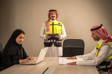 دراسة الرسوم البيانية والاحصاءات، متابعة مخطط الاعمار, العمل في القطاع الهندسي، مفهوم الهندسة والانشاءات المعمارية، مهندس عربي خليجي سعودي يرتدي الثوب التقليدي والشماغ وسترة الحماية يمسك بيده المخطط يجلس مع موظفة تمسك بيدها جهاز اللابتوب ومهندس معماري  ، مهنة ووظيفة في السعودية