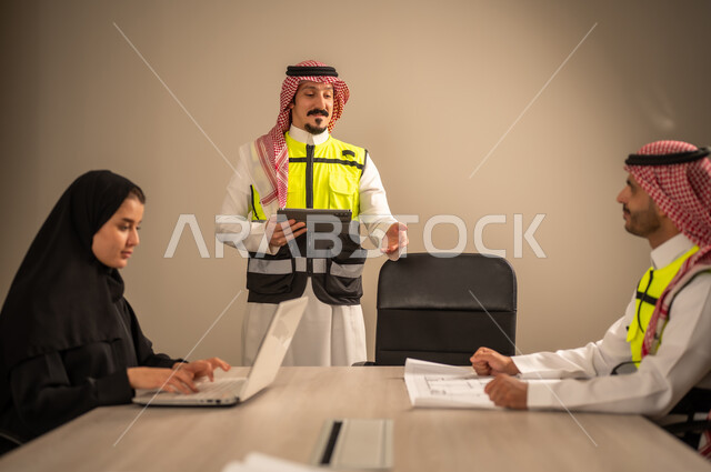 دراسة الرسوم البيانية والاحصاءات، متابعة مخطط الاعمار, العمل في القطاع الهندسي، مفهوم الهندسة والانشاءات المعمارية، مهندس عربي خليجي سعودي يرتدي الثوب التقليدي والشماغ وسترة الحماية يمسك بيده المخطط يجلس مع موظفة تمسك بيدها جهاز اللابتوب ومهندس معماري  ، مهنة ووظيفة في السعودية