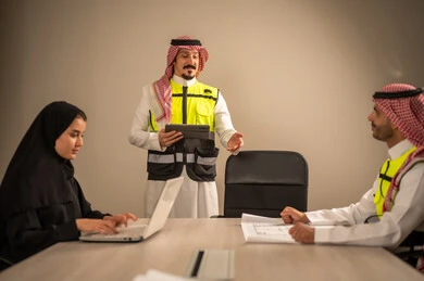 دراسة الرسوم البيانية والاحصاءات، متابعة مخطط الاعمار, العمل في القطاع الهندسي، مفهوم الهندسة والانشاءات المعمارية، مهندس عربي خليجي سعودي يرتدي الثوب التقليدي والشماغ وسترة الحماية يمسك بيده المخطط يجلس مع موظفة تمسك بيدها جهاز اللابتوب ومهندس معماري  ، مهنة ووظيفة في السعودية