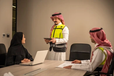 دراسة الرسوم البيانية والاحصاءات، متابعة مخطط الاعمار, العمل في القطاع الهندسي، مفهوم الهندسة والانشاءات المعمارية، مهندس عربي خليجي سعودي يرتدي الثوب التقليدي والشماغ وسترة الحماية يمسك بيده المخطط يجلس مع موظفة تمسك بيدها جهاز اللابتوب ومهندس معماري  ، مهنة ووظيفة في السعودية