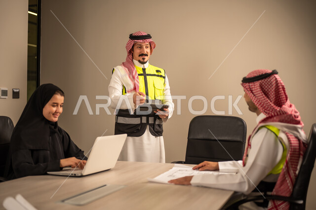 دراسة الرسوم البيانية والاحصاءات، متابعة مخطط الاعمار, العمل في القطاع الهندسي، مفهوم الهندسة والانشاءات المعمارية، مهندس عربي خليجي سعودي يرتدي الثوب التقليدي والشماغ وسترة الحماية يمسك بيده المخطط يجلس مع موظفة تمسك بيدها جهاز اللابتوب ومهندس معماري  ، مهنة ووظيفة في السعودية