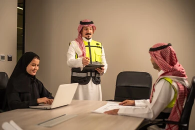 دراسة الرسوم البيانية والاحصاءات، متابعة مخطط الاعمار, العمل في القطاع الهندسي، مفهوم الهندسة والانشاءات المعمارية، مهندس عربي خليجي سعودي يرتدي الثوب التقليدي والشماغ وسترة الحماية يمسك بيده المخطط يجلس مع موظفة تمسك بيدها جهاز اللابتوب ومهندس معماري  ، مهنة ووظيفة في السعودية