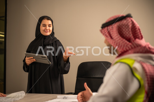 دراسة الرسوم البيانية والاحصاءات، متابعة مخطط الاعمار, العمل في القطاع الهندسي، مفهوم الهندسة والانشاءات المعمارية، موظفة سعودية تمسك بيدها جهاز التابلت تتناقش مع مهندسين عربيين خليجيين سعوديين يرتديون الثوب التقليدي والشماغ وسترة الحماية امامهم المخطط  ، مهنة ووظيفة في السعودية