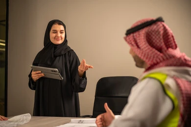 دراسة الرسوم البيانية والاحصاءات، متابعة مخطط الاعمار, العمل في القطاع الهندسي، مفهوم الهندسة والانشاءات المعمارية، موظفة سعودية تمسك بيدها جهاز التابلت تتناقش مع مهندسين عربيين خليجيين سعوديين يرتديون الثوب التقليدي والشماغ وسترة الحماية امامهم المخطط  ، مهنة ووظيفة في السعودية