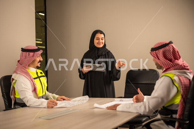 دراسة الرسوم البيانية والاحصاءات، متابعة مخطط الاعمار, العمل في القطاع الهندسي، مفهوم الهندسة والانشاءات المعمارية، موظفة سعودية تمسك بيدها جهاز التابلت تتناقش مع مهندسين عربيين خليجيين سعوديين يرتديون الثوب التقليدي والشماغ وسترة الحماية امامهم المخطط  ، مهنة ووظيفة في السعودية
