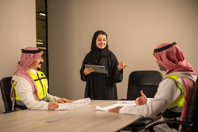 دراسة الرسوم البيانية والاحصاءات، متابعة مخطط الاعمار, العمل في القطاع الهندسي، مفهوم الهندسة والانشاءات المعمارية، موظفة سعودية تمسك بيدها جهاز التابلت تتناقش مع مهندسين عربيين خليجيين سعوديين يرتديون الثوب التقليدي والشماغ وسترة الحماية امامهم المخطط  ، مهنة ووظيفة في السعودية