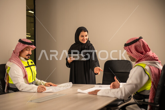 دراسة الرسوم البيانية والاحصاءات، متابعة مخطط الاعمار, العمل في القطاع الهندسي، مفهوم الهندسة والانشاءات المعمارية، موظفة سعودية تمسك بيدها جهاز التابلت تتناقش مع مهندسين عربيين خليجيين سعوديين يرتديون الثوب التقليدي والشماغ وسترة الحماية امامهم المخطط  ، مهنة ووظيفة في السعودية
