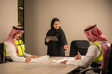 دراسة الرسوم البيانية والاحصاءات، متابعة مخطط الاعمار, العمل في القطاع الهندسي، مفهوم الهندسة والانشاءات المعمارية، موظفة سعودية تمسك بيدها جهاز التابلت تتناقش مع مهندسين عربيين خليجيين سعوديين يرتديون الثوب التقليدي والشماغ وسترة الحماية امامهم المخطط  ، مهنة ووظيفة في السعودية