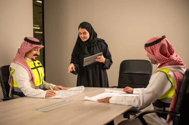 دراسة الرسوم البيانية والاحصاءات، متابعة مخطط الاعمار, العمل في القطاع الهندسي، مفهوم الهندسة والانشاءات المعمارية، موظفة سعودية تمسك بيدها جهاز التابلت تتناقش مع مهندسين عربيين خليجيين سعوديين يرتديون الثوب التقليدي والشماغ وسترة الحماية امامهم المخطط  ، مهنة ووظيفة في السعودية