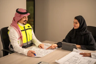 دراسة الرسوم البيانية والاحصاءات، متابعة مخطط الاعمار, العمل في القطاع الهندسي، مفهوم الهندسة والانشاءات المعمارية، موظفة سعودية تمسك بيدها جهاز التابلت تتناقش مع مهندسين عربيين خليجيين سعوديين يرتديون الثوب التقليدي والشماغ وسترة الحماية امامهم المخطط  ، مهنة ووظيفة في السعودية