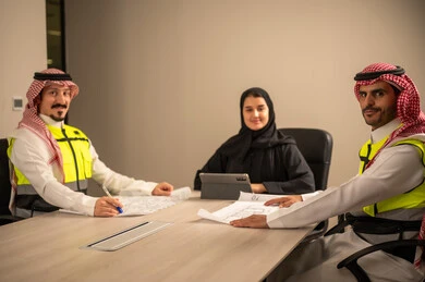 دراسة الرسوم البيانية والاحصاءات، متابعة مخطط الاعمار, العمل في القطاع الهندسي، مفهوم الهندسة والانشاءات المعمارية، موظفة سعودية تمسك بيدها جهاز التابلت تتناقش مع مهندسين عربيين خليجيين سعوديين يرتديون الثوب التقليدي والشماغ وسترة الحماية امامهم المخطط  ، مهنة ووظيفة في السعودية