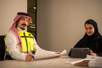 دراسة الرسوم البيانية والاحصاءات، متابعة مخطط الاعمار, العمل في القطاع الهندسي، مفهوم الهندسة والانشاءات المعمارية، موظفة سعودية تمسك بيدها جهاز التابلت تتناقش مع مهندسين عربيين خليجيين سعوديين يرتديون الثوب التقليدي والشماغ وسترة الحماية امامهم المخطط  ، مهنة ووظيفة في السعودية