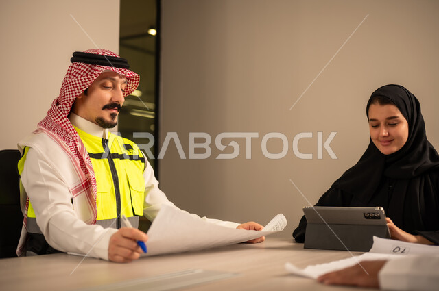 دراسة الرسوم البيانية والاحصاءات، متابعة مخطط الاعمار, العمل في القطاع الهندسي، مفهوم الهندسة والانشاءات المعمارية، مهندس عربي خليجي سعودي يرتدي الثوب التقليدي والشماغ وسترة الحماية يمسك بيده المخطط يجلس مع موظفة تمسك بيدها جهاز التابلت ، مهنة ووظيفة في السعودية