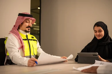 دراسة الرسوم البيانية والاحصاءات، متابعة مخطط الاعمار, العمل في القطاع الهندسي، مفهوم الهندسة والانشاءات المعمارية، مهندس عربي خليجي سعودي يرتدي الثوب التقليدي والشماغ وسترة الحماية يمسك بيده المخطط يجلس مع موظفة تمسك بيدها جهاز التابلت ، مهنة ووظيفة في السعودية