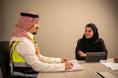 دراسة الرسوم البيانية والاحصاءات، متابعة مخطط الاعمار, العمل في القطاع الهندسي، مفهوم الهندسة والانشاءات المعمارية، مهندس عربي خليجي سعودي يرتدي الثوب التقليدي والشماغ وسترة الحماية يمسك بيده المخطط يجلس مع موظفة تمسك بيدها جهاز التابلت ، مهنة ووظيفة في السعودية