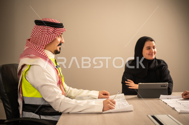 دراسة الرسوم البيانية والاحصاءات، متابعة مخطط الاعمار, العمل في القطاع الهندسي، مفهوم الهندسة والانشاءات المعمارية، مهندس عربي خليجي سعودي يرتدي الثوب التقليدي والشماغ وسترة الحماية يمسك بيده المخطط يجلس مع موظفة تمسك بيدها جهاز التابلت ، مهنة ووظيفة في السعودية