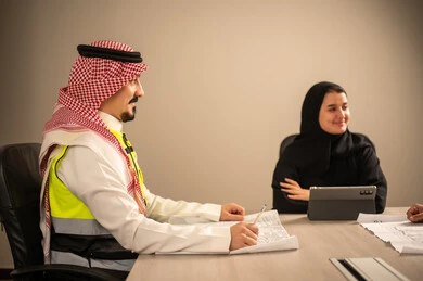 Studying charts and statistics, following up on the construction plan, working in the engineering sector, the concept of engineering and architectural constructions, a Saudi Gulf Arab engineer wearing traditional attire and a headscarf, holding a plan in his hand, sitting with a female employee holding a tablet in her hand, profession and job in Saudi Arabia.