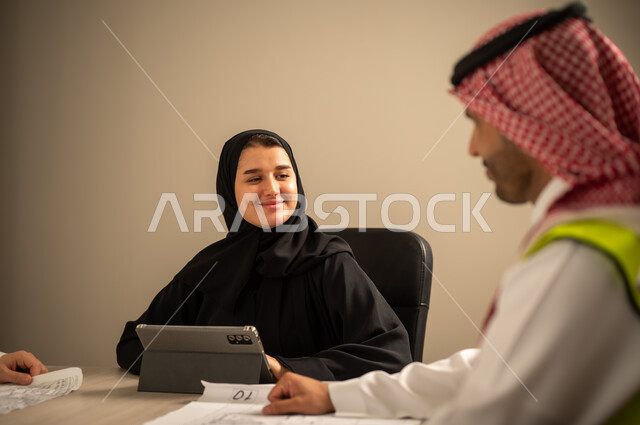 دراسة الرسوم البيانية والاحصاءات، متابعة مخطط الاعمار, العمل في القطاع الهندسي، مفهوم الهندسة والانشاءات المعمارية، مهندس عربي خليجي سعودي يرتدي الثوب التقليدي والشماغ وسترة الحماية يمسك بيده المخطط يجلس مع موظفة تمسك بيدها جهاز التابلت ، مهنة ووظيفة في السعودية