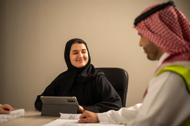 دراسة الرسوم البيانية والاحصاءات، متابعة مخطط الاعمار, العمل في القطاع الهندسي، مفهوم الهندسة والانشاءات المعمارية، مهندس عربي خليجي سعودي يرتدي الثوب التقليدي والشماغ وسترة الحماية يمسك بيده المخطط يجلس مع موظفة تمسك بيدها جهاز التابلت ، مهنة ووظيفة في السعودية