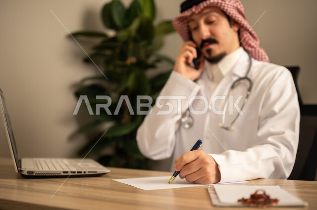 استعمال الهاتف المحمول، قراءة التحاليل والتقارير الطبية، طبيب عربي خليجي سعودي يرتدي المعطف الأبيض يضع السماعات الطبيه حول رقبته يستخدم الحاسوب المحمول، يحمل بيده أوراق وملفات المرضى، العمل في القطاع الصحي في المملكة العربية السعودية، تقديم الرعاية الصحية للمرضى