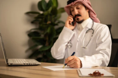 استعمال الهاتف المحمول، قراءة التحاليل والتقارير الطبية، طبيب عربي خليجي سعودي يرتدي المعطف الأبيض يضع السماعات الطبيه حول رقبته يستخدم الحاسوب المحمول، يحمل بيده أوراق وملفات المرضى، العمل في القطاع الصحي في المملكة العربية السعودية، تقديم الرعاية الصحية للمرضى