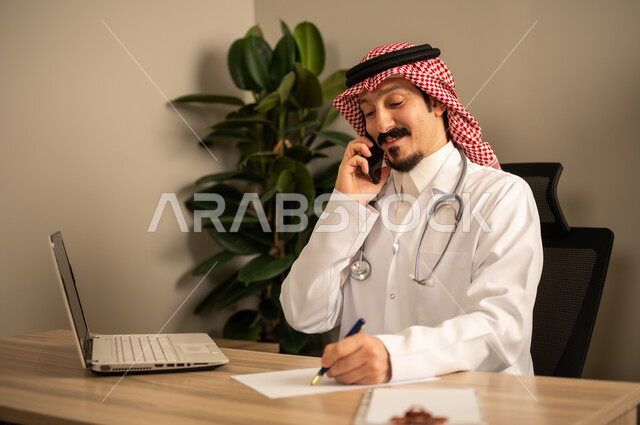 استعمال الهاتف المحمول، قراءة التحاليل والتقارير الطبية، طبيب عربي خليجي سعودي يرتدي المعطف الأبيض يضع السماعات الطبيه حول رقبته يستخدم الحاسوب المحمول، يحمل بيده أوراق وملفات المرضى، العمل في القطاع الصحي في المملكة العربية السعودية، تقديم الرعاية الصحية للمرضى