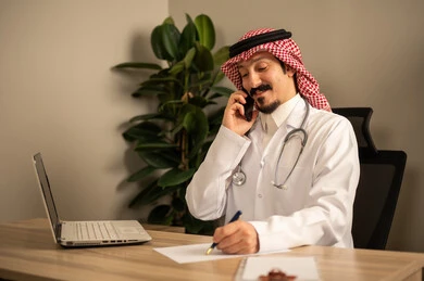 استعمال الهاتف المحمول، قراءة التحاليل والتقارير الطبية، طبيب عربي خليجي سعودي يرتدي المعطف الأبيض يضع السماعات الطبيه حول رقبته يستخدم الحاسوب المحمول، يحمل بيده أوراق وملفات المرضى، العمل في القطاع الصحي في المملكة العربية السعودية، تقديم الرعاية الصحية للمرضى