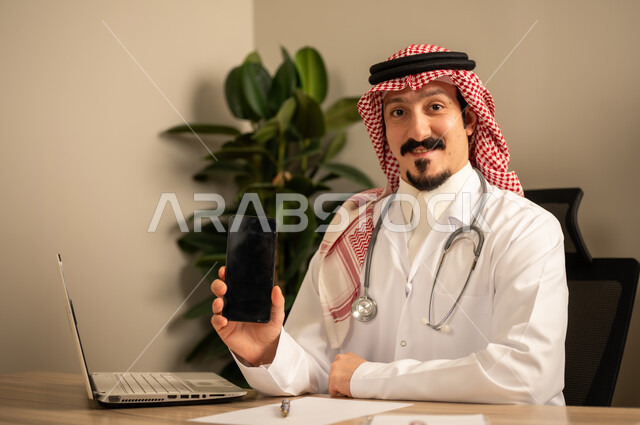 استعمال الهاتف المحمول، قراءة التحاليل والتقارير الطبية، طبيب عربي خليجي سعودي يرتدي المعطف الأبيض يضع السماعات الطبيه حول رقبته يستخدم الحاسوب المحمول، يحمل بيده أوراق وملفات المرضى، العمل في القطاع الصحي في المملكة العربية السعودية، تقديم الرعاية الصحية للمرضى