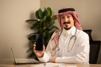 استعمال الهاتف المحمول، قراءة التحاليل والتقارير الطبية، طبيب عربي خليجي سعودي يرتدي المعطف الأبيض يضع السماعات الطبيه حول رقبته يستخدم الحاسوب المحمول، يحمل بيده أوراق وملفات المرضى، العمل في القطاع الصحي في المملكة العربية السعودية، تقديم الرعاية الصحية للمرضى