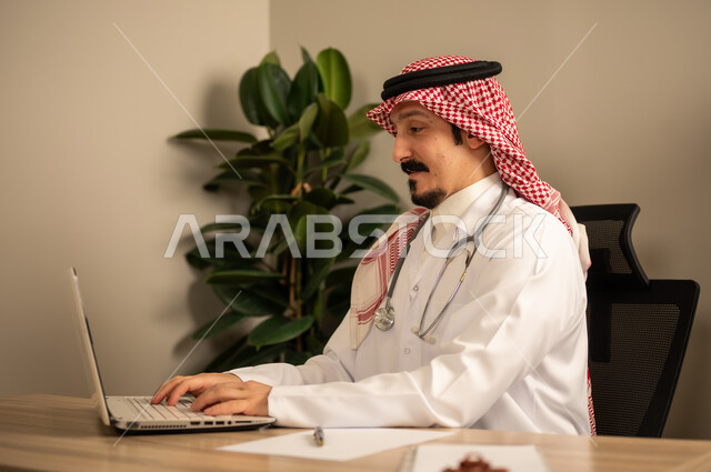 قراءة التحاليل والتقارير الطبية، طبيب عربي خليجي سعودي يرتدي المعطف الأبيض يضع السماعات الطبيه حول رقبته يستخدم الحاسوب المحمول، يحمل بيده أوراق وملفات المرضى، العمل في القطاع الصحي في المملكة العربية السعودية، تقديم الرعاية الصحية للمرضى