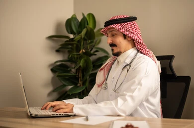 قراءة التحاليل والتقارير الطبية، طبيب عربي خليجي سعودي يرتدي المعطف الأبيض يضع السماعات الطبيه حول رقبته يستخدم الحاسوب المحمول، يحمل بيده أوراق وملفات المرضى، العمل في القطاع الصحي في المملكة العربية السعودية، تقديم الرعاية الصحية للمرضى