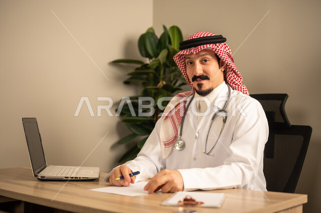 قراءة التحاليل والتقارير الطبية، طبيب عربي خليجي سعودي يرتدي المعطف الأبيض يضع السماعات الطبيه حول رقبته يستخدم الحاسوب المحمول، يحمل بيده أوراق وملفات المرضى، العمل في القطاع الصحي في المملكة العربية السعودية، تقديم الرعاية الصحية للمرضى