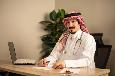 Reading medical analyses and reports, an Arab Gulf Saudi doctor wearing a white coat with a stethoscope around his neck using a laptop, holding patient papers and files in his hand, working in the health sector in the Kingdom of Saudi Arabia, providing healthcare to patients.